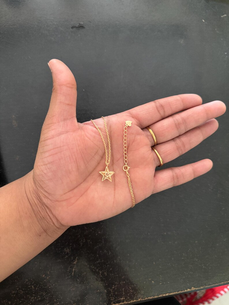 A gold necklace with a star pendant and an adjustable chain extension, displayed on an open hand alongside two gold rings against a dark gray background.