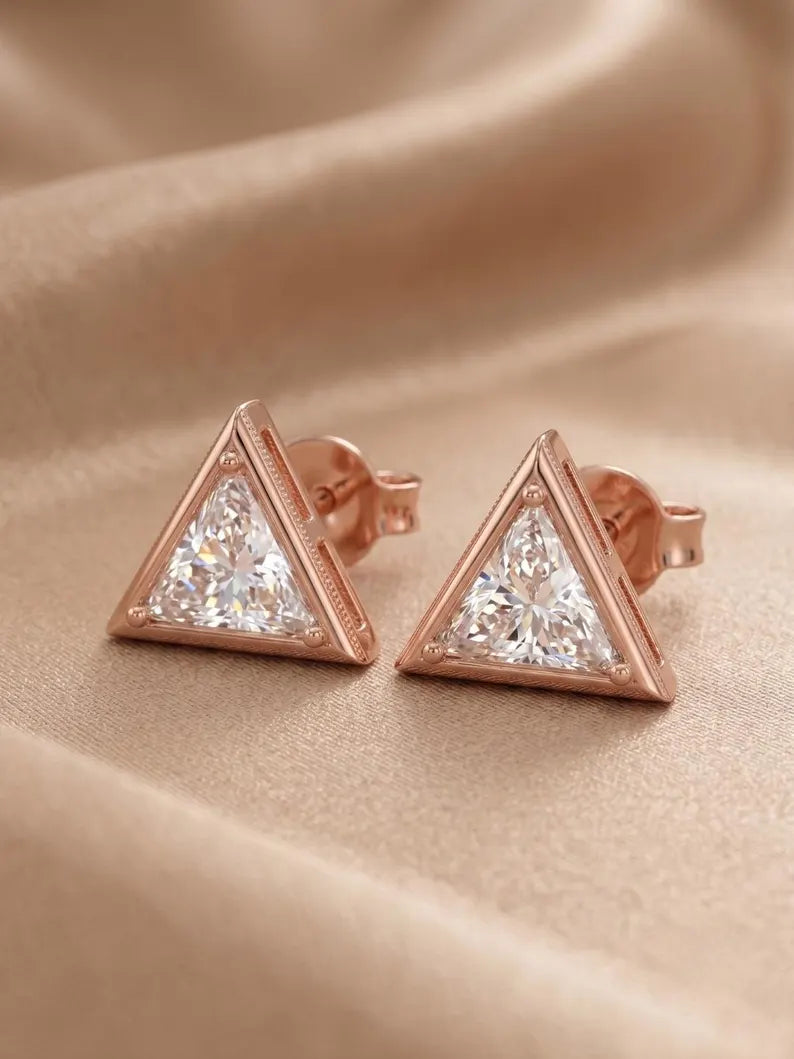 A pair of rose gold triangle stud earrings with clear faceted gemstones, displayed on a draped beige satin fabric background.