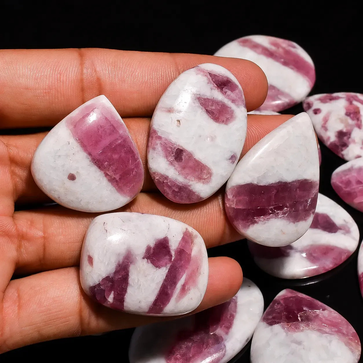 Polished rhodonite stones with pink and white bands held in a hand against a dark background.