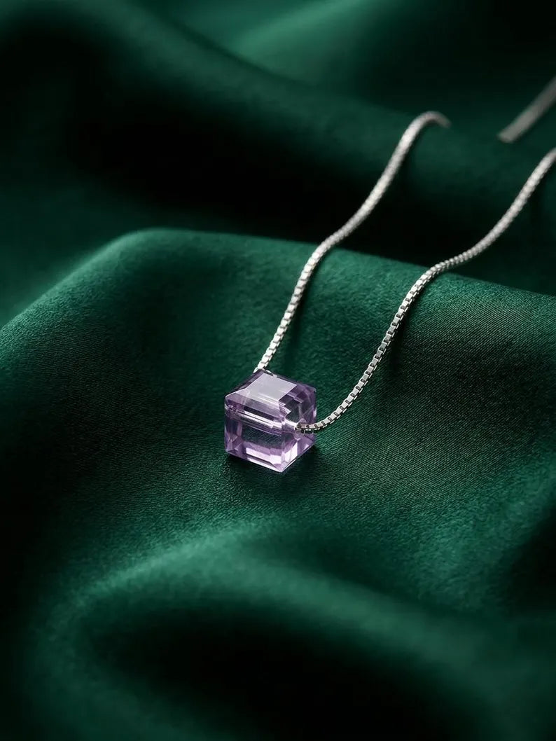 A silver chain necklace with a faceted purple gemstone pendant, displayed on draped dark green velvet fabric.