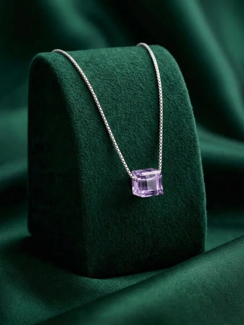A silver chain necklace with a faceted purple gemstone pendant, displayed on a dark green velvet jewelry stand against a matching green fabric background.