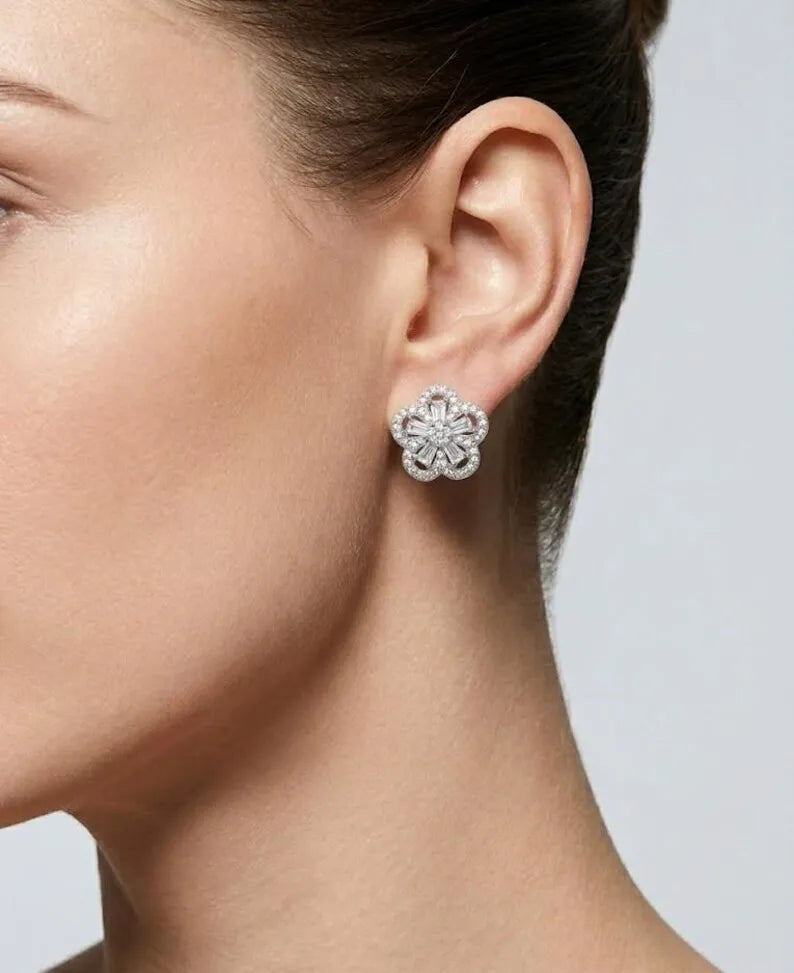 A close-up side profile of a woman's ear wearing a small flower-shaped stud earring with sparkling clear stones in a silver-toned setting, against a light gray background.