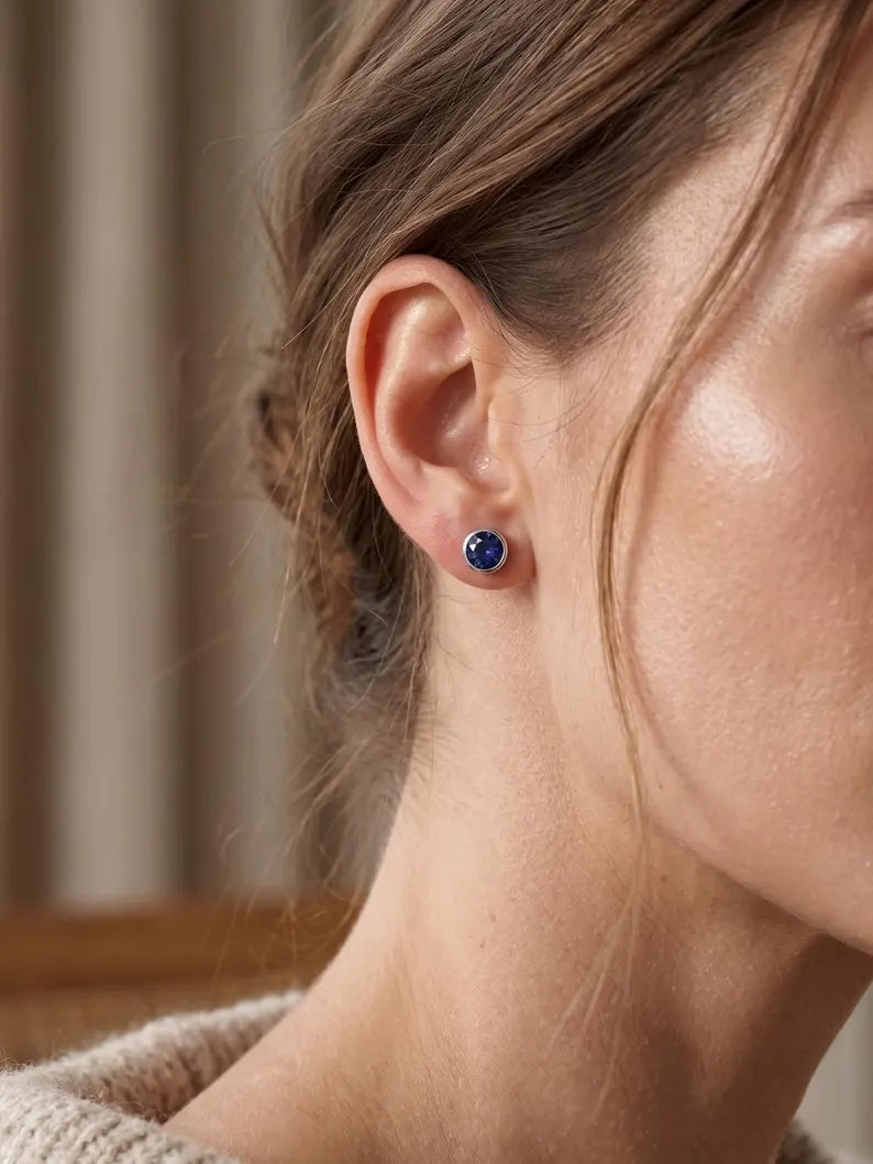 A close-up of a woman's ear wearing a small round stud earring with a deep blue stone in a silver setting, against a softly blurred indoor background.