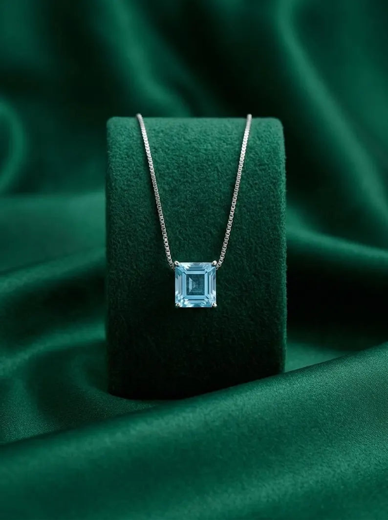 A silver necklace with an emerald-cut light blue gemstone pendant, displayed on a dark green velvet jewelry stand against a matching green satin fabric background.