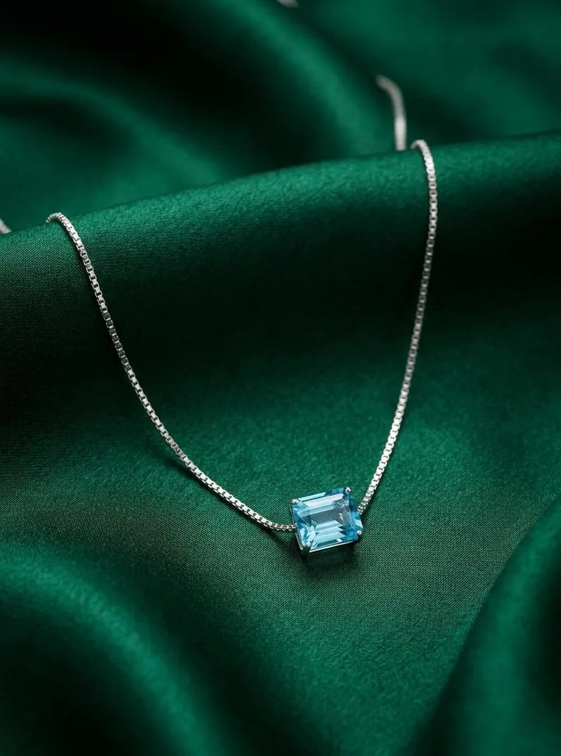 A silver chain necklace with a rectangular light blue gemstone pendant, displayed on draped emerald green satin fabric.