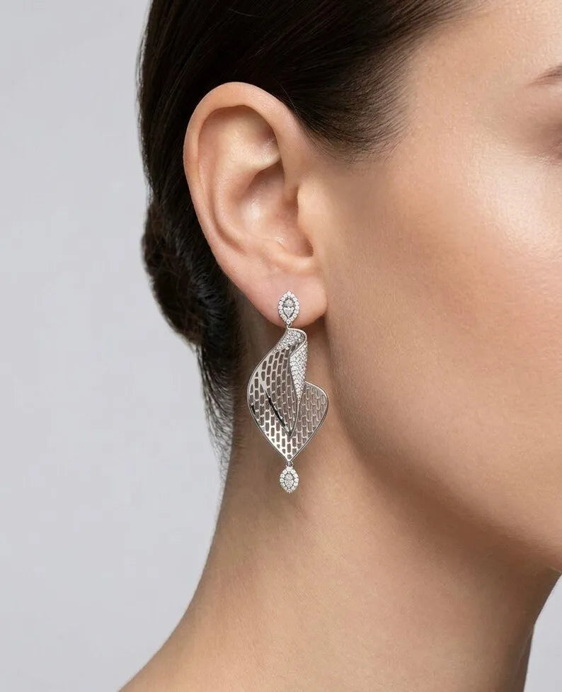 A close-up of a model's ear wearing a silver-toned, leaf-shaped drop earring adorned with small clear stones.