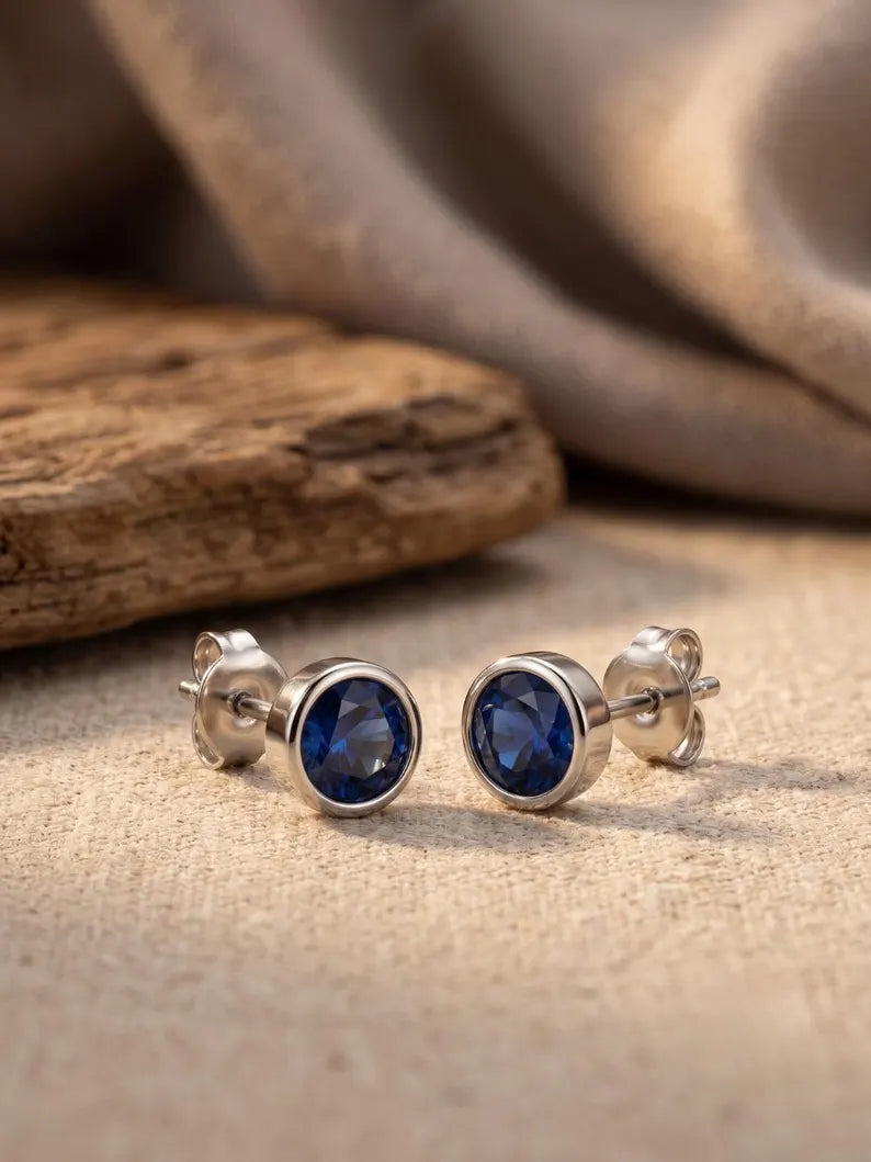 A pair of silver stud earrings with oval deep blue gemstones, displayed on a textured beige fabric surface next to a piece of rustic wood.