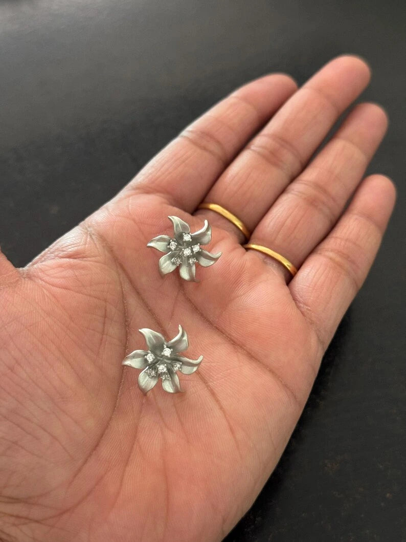 A close-up of a person's hand holding two small silver flower-shaped earrings, with two gold rings on the fingers, against a dark background.