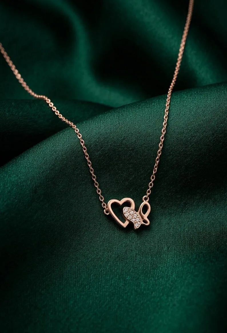A rose gold necklace with a double-heart pendant, one of which is adorned with small clear crystals, displayed on a draped dark green satin fabric.