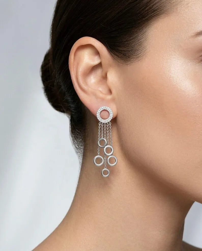 A close-up of a model's ear wearing a silver dangle earring with a circular stud and three cascading chain loops.