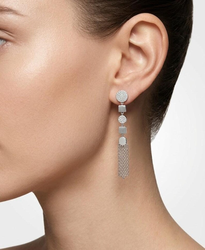 A close-up of a model's ear wearing a long silver dangle earring with a round pavé stud and a cascading drop of small silver and white stones ending in a textured fringe.