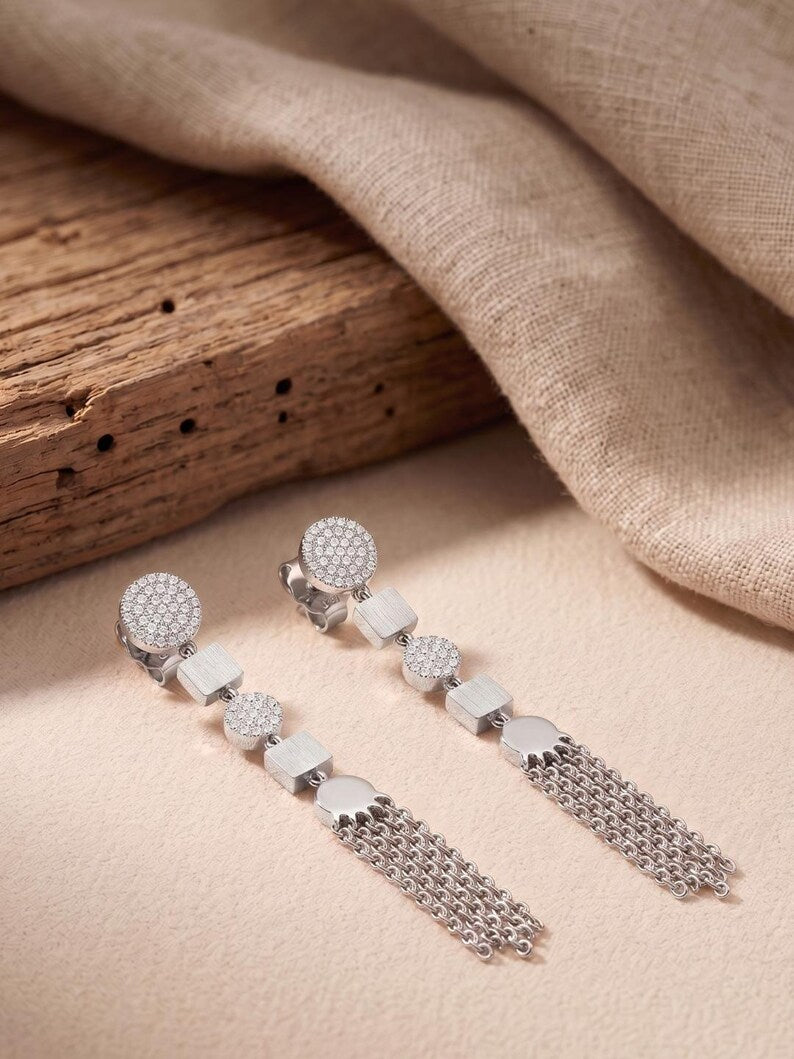 A pair of silver-toned statement dangle earrings with round pavé-set stones, square links, and dangling chain fringe, styled on a beige fabric surface with a rustic wood block and draped textile in the background.