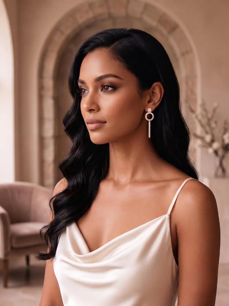 A woman with long, wavy black hair wearing a cream satin slip dress and a long silver key-shaped dangle earring, posed in front of a warm beige stone archway background.