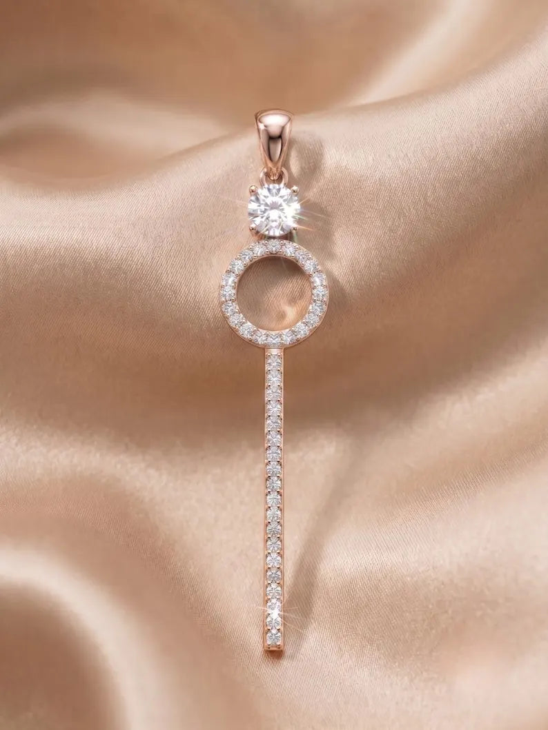 A rose gold key-shaped pendant with a round clear gemstone at the top and a vertical bar of small clear stones, displayed on a beige satin fabric background.