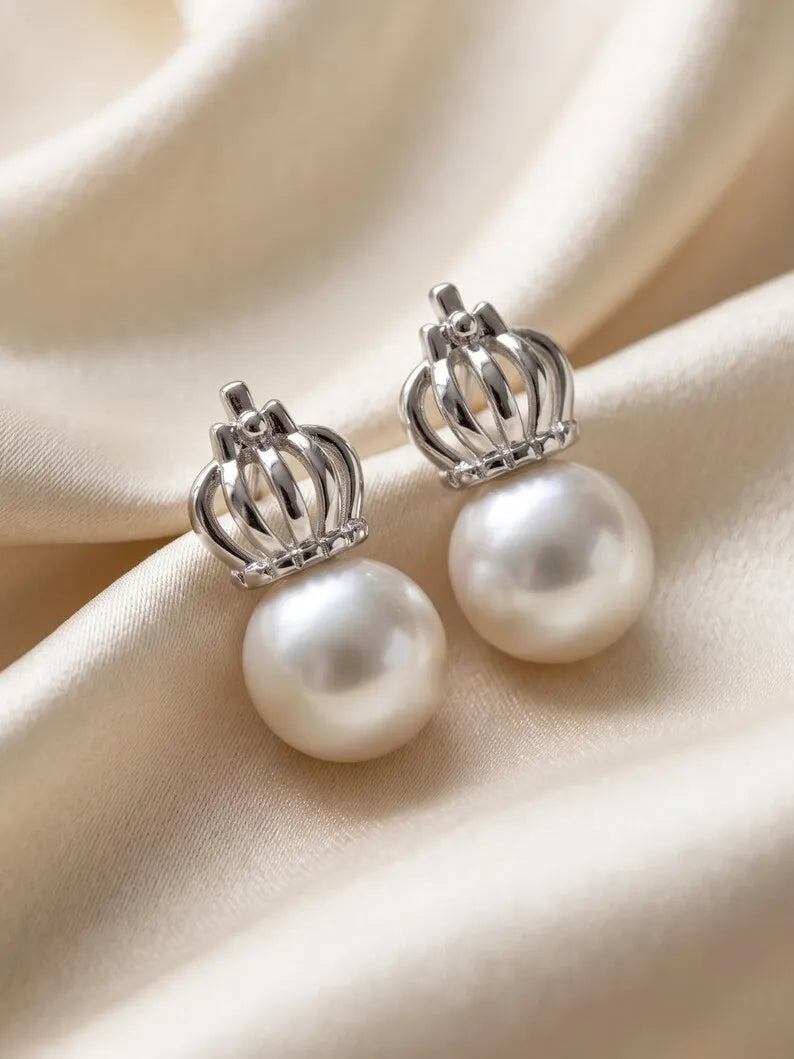 A pair of crown-shaped silver stud earrings with large white pearl drops, displayed on draped cream satin fabric.