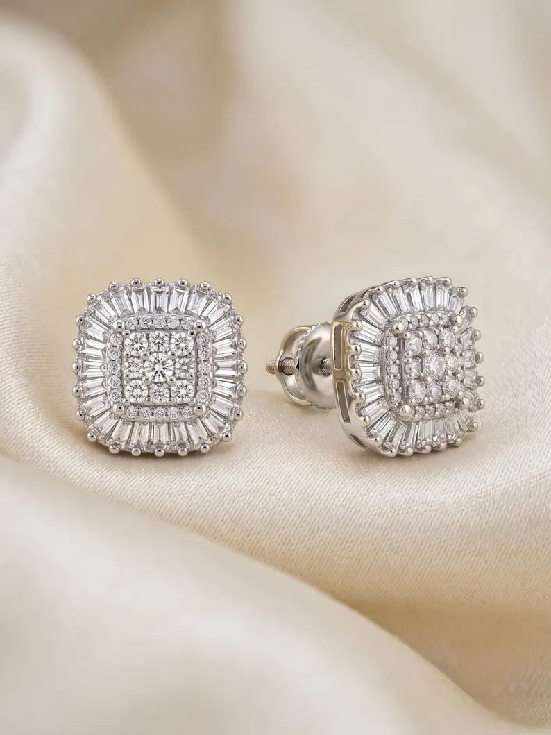 A pair of square stud earrings with a silver-toned halo setting and clear baguette and round stones, displayed on a draped cream satin fabric.