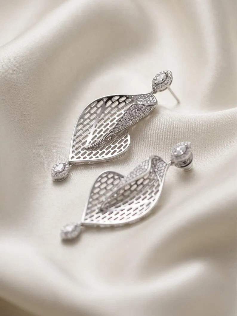 A pair of silver drop earrings with leaf-shaped filigree pendants and small oval clear stones, displayed on draped cream satin fabric.