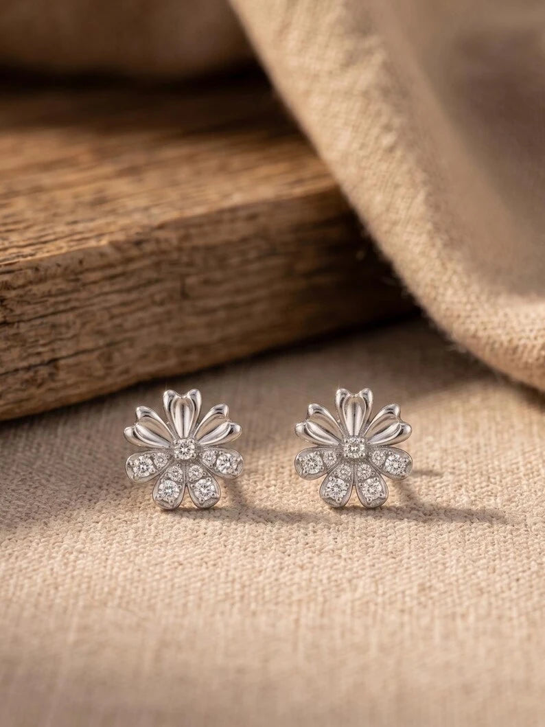 A pair of silver-toned flower-shaped stud earrings with clear stones, displayed on a beige fabric surface next to a wooden block.