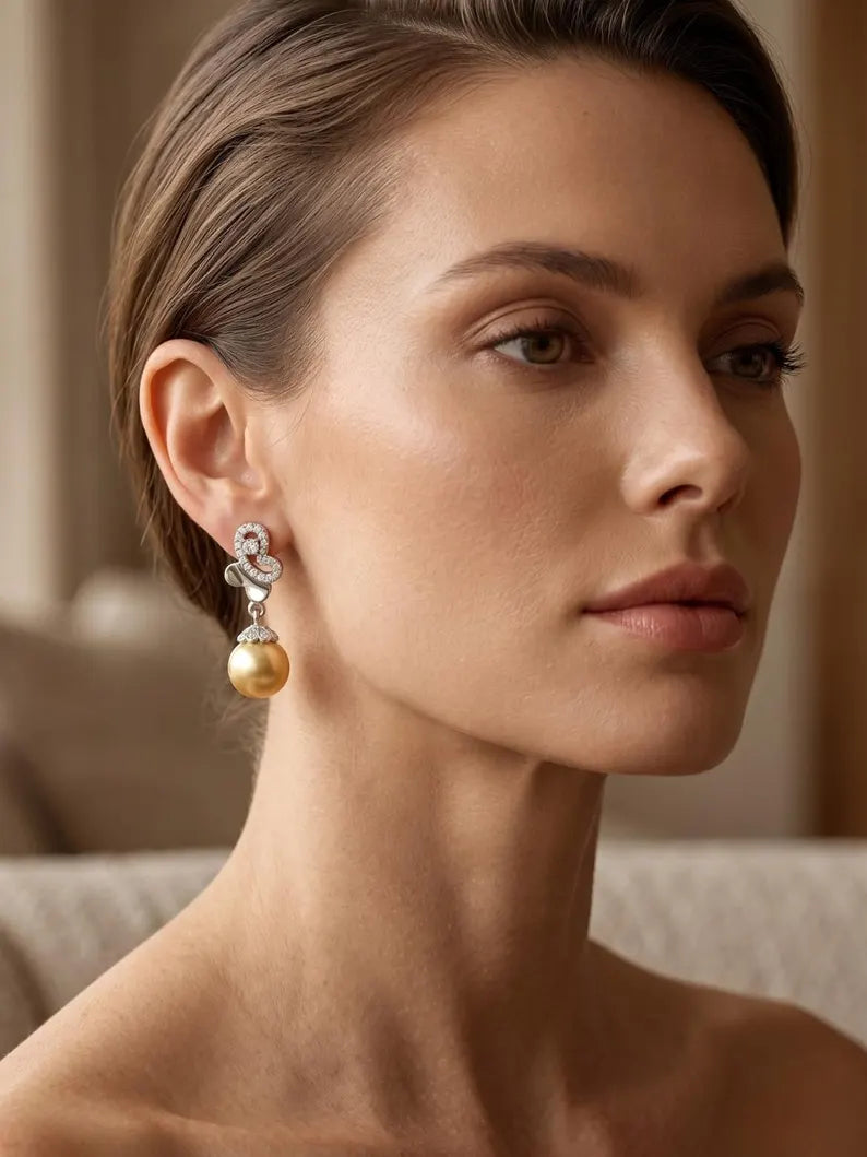 A close-up portrait of a woman wearing a dangling earring with a gold pearl and a silver-toned floral scroll setting with clear stones.