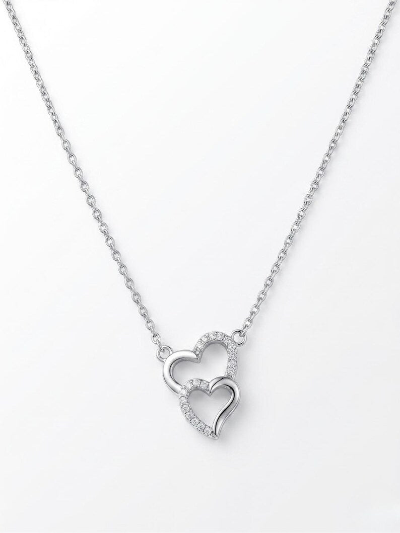 A silver-toned chain necklace featuring two interlocking open heart pendants, one adorned with small clear stones, on a plain white background.