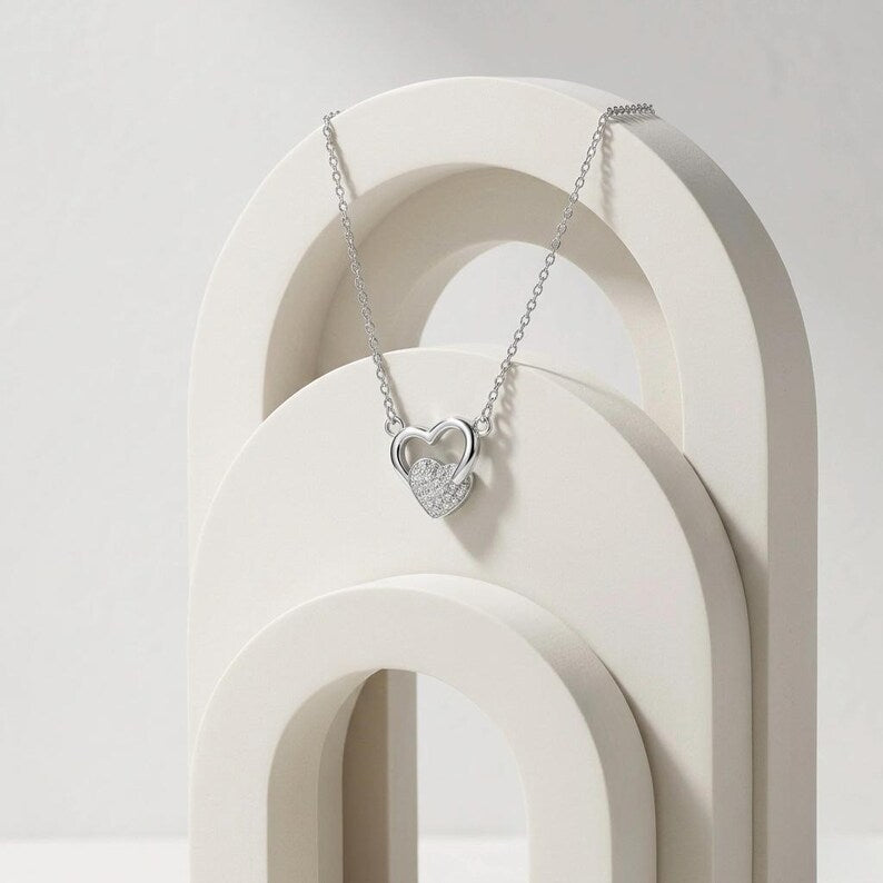 A silver heart pendant necklace with a pavé-set clear stone center, draped over a white arched display stand against a light gray background.