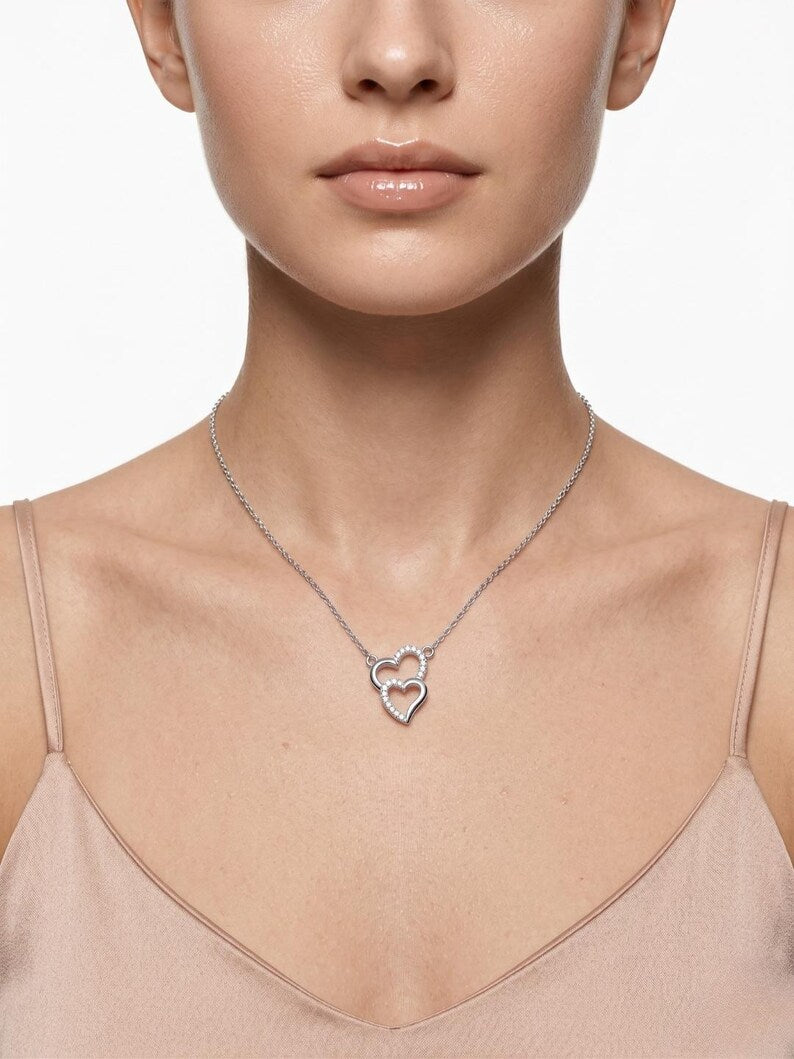 A close-up of a model wearing a delicate silver chain necklace with a double interlocking heart pendant, styled with a beige camisole top against a clean white background.