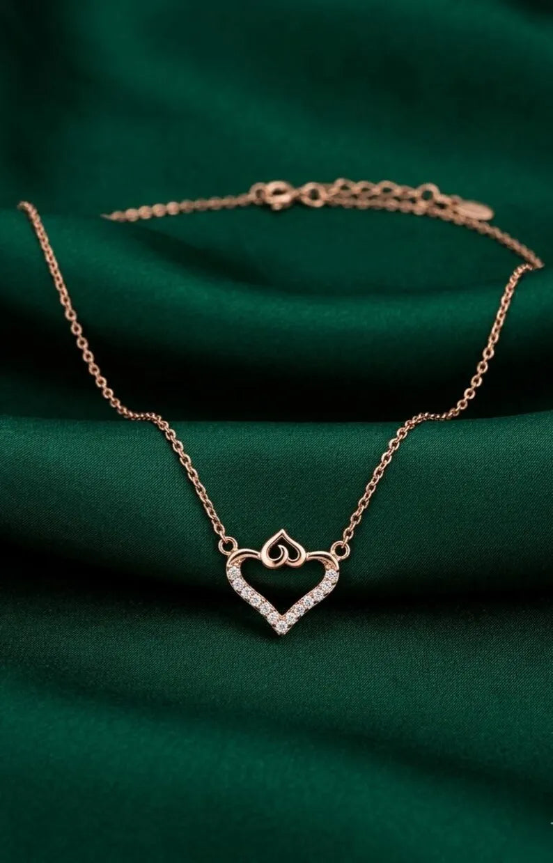 A rose gold necklace featuring a double-heart pendant with a border of small clear stones, displayed on a draped dark green fabric background.
