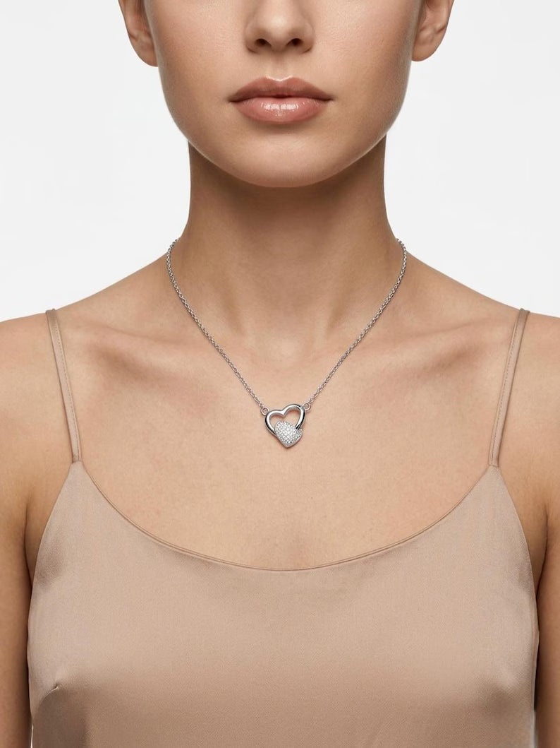 A close-up of a model wearing a delicate silver chain necklace with a small, open heart pendant set with tiny clear stones, paired with a beige camisole top against a light gray background.