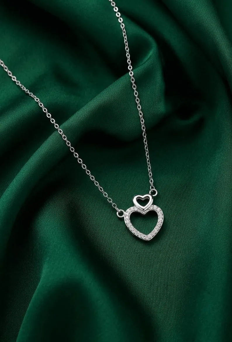 A silver double-heart pendant necklace with a pavé-set heart and a smaller hollow heart above it, displayed on draped dark green satin fabric.