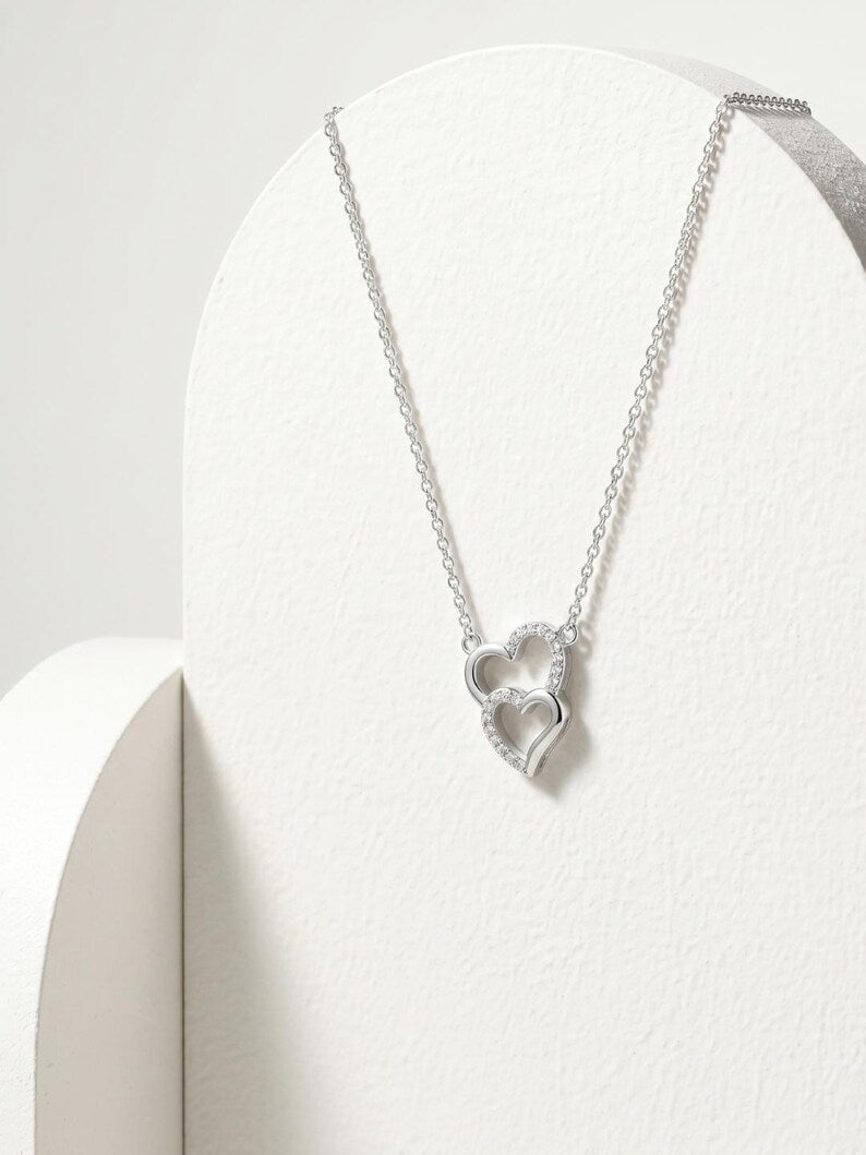 A silver necklace with a double interlocking heart pendant, displayed on a white curved jewelry stand against a light gray background.