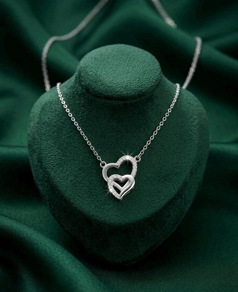 A silver necklace with a double interlocking heart pendant, displayed on a dark green velvet jewelry bust against a matching green fabric background.