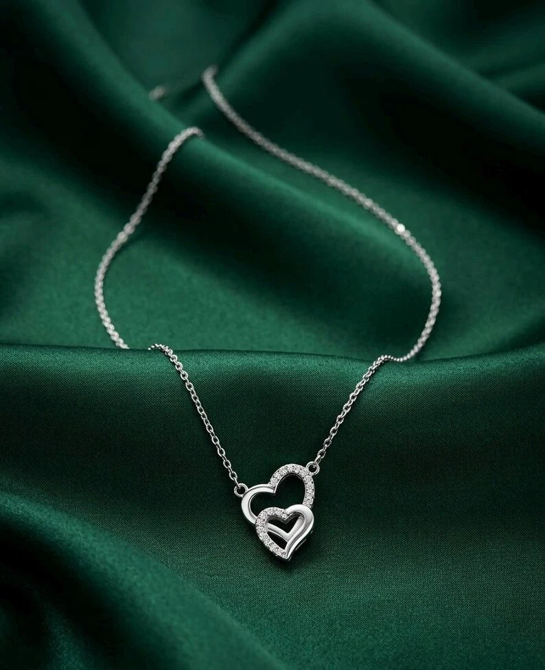 A silver double-heart pendant necklace with sparkling clear stones, resting on draped dark green satin fabric.