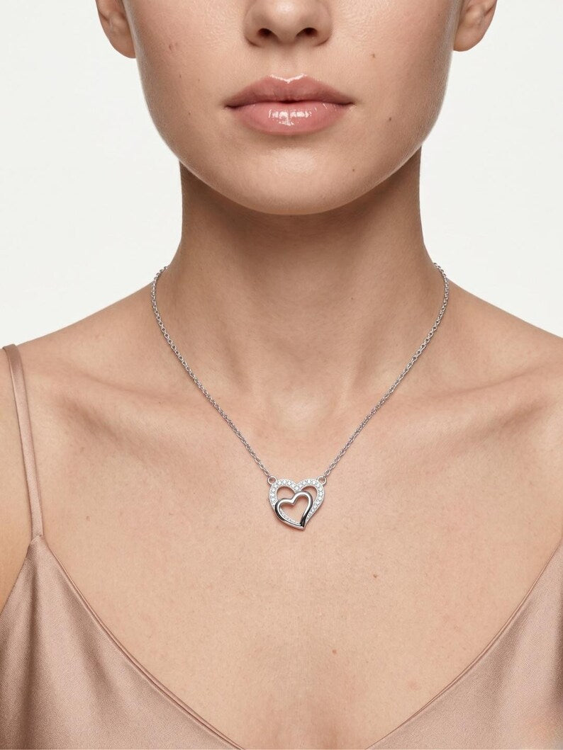 A close-up of a model wearing a delicate silver chain necklace with a double-heart pendant, one heart inlaid with small clear stones and the other featuring a small red heart in the center.