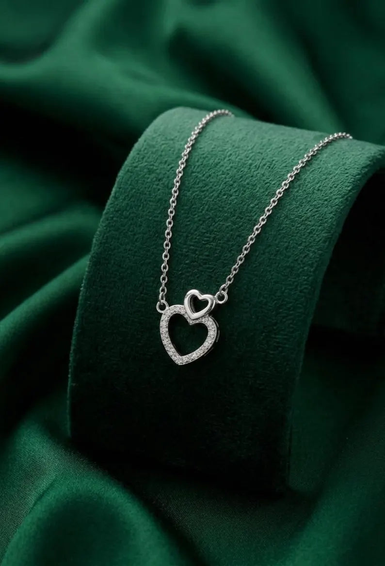 A silver necklace with a double-heart pendant, displayed on a dark green velvet jewelry stand against a matching green fabric background.