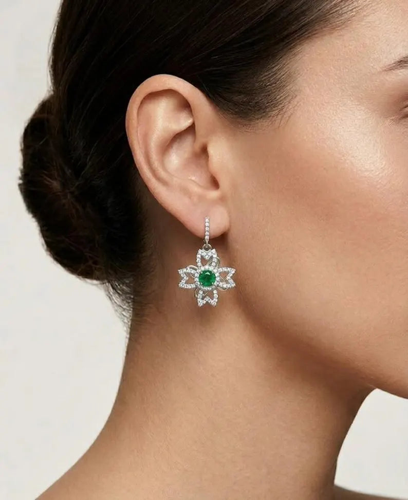 A close-up of a model's ear wearing a silver-toned dangle earring with a green emerald center and clear stones in a floral starburst design.