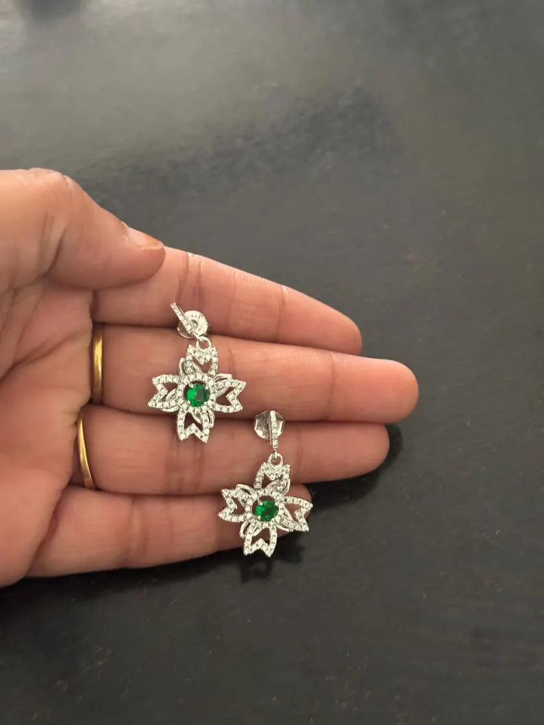 A pair of silver-toned dangle earrings with green center stones and clear surrounding stones, held in a person's hand against a dark gray background.