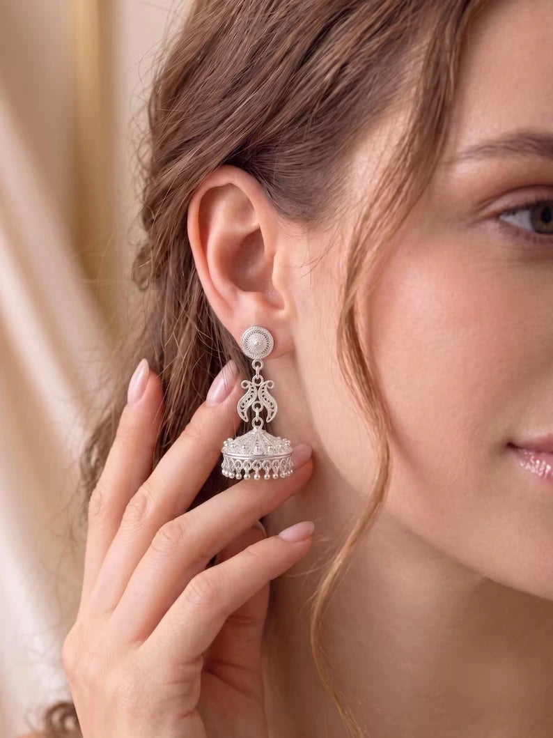 A close-up of a woman wearing a silver-toned traditional Indian jhumka earring with a white pearl-like round stud and a dangling ornate bell-shaped charm.