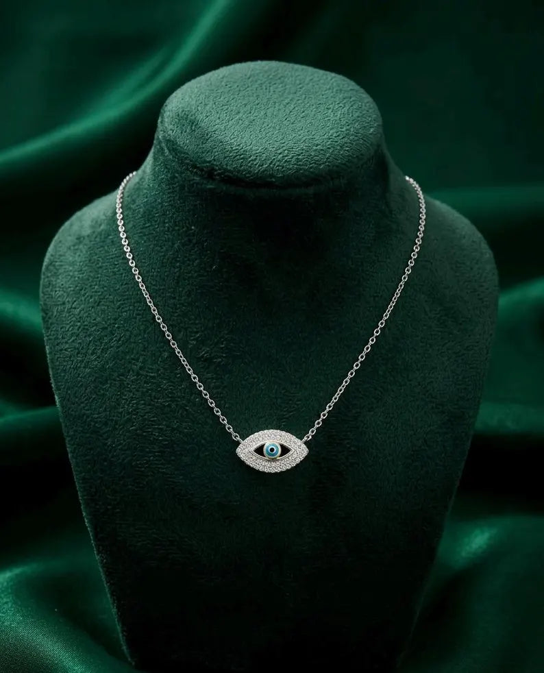 A silver-toned evil eye necklace with a turquoise-blue center stone, displayed on a dark green velvet jewelry bust against a matching green satin background.