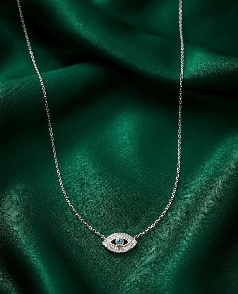 A silver-toned evil eye pendant necklace with a small green center stone, displayed on draped emerald green satin fabric.