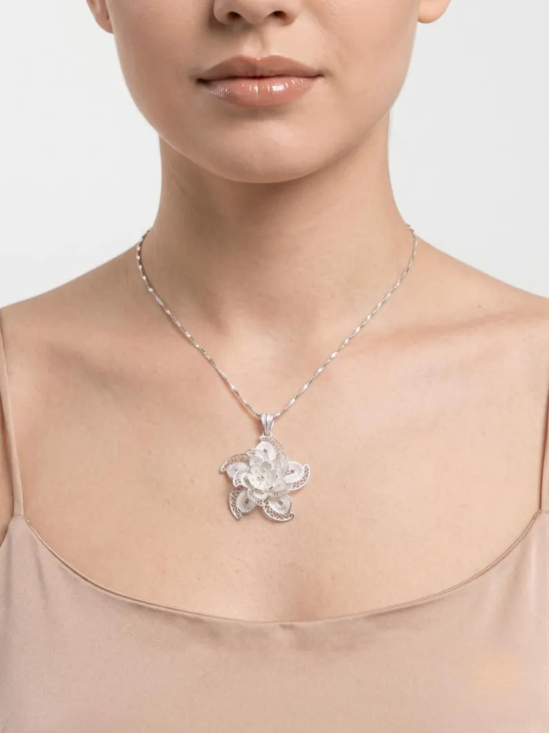 A close-up of a model wearing a delicate silver chain necklace with a silver-toned flower-shaped pendant, paired with a beige satin top against a light gray background.