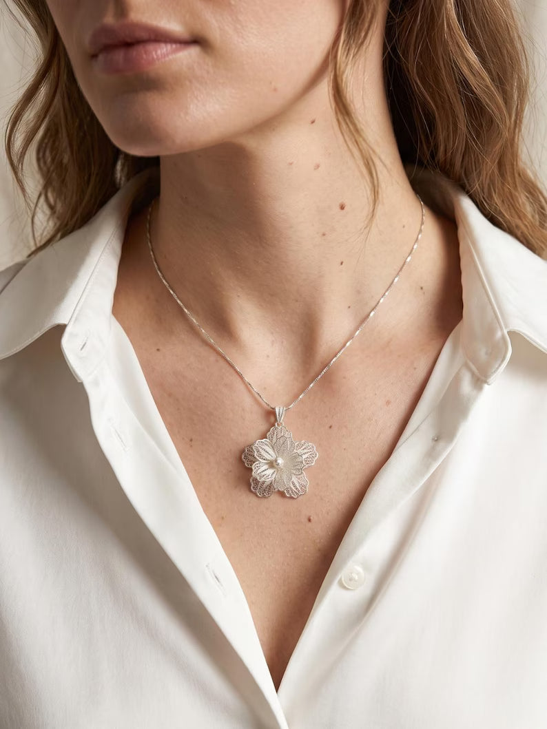 A close-up of a model wearing a delicate silver flower pendant necklace with a textured silver chain, styled with a white button-down shirt.