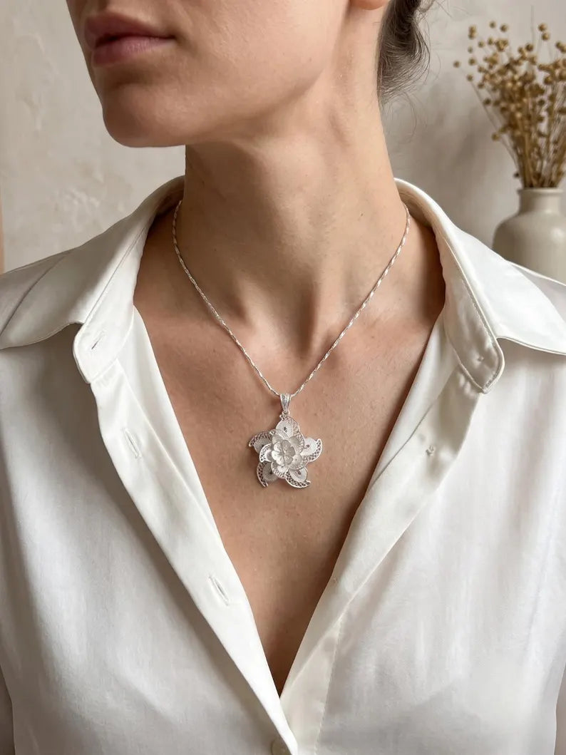 A close-up of a woman wearing a silver floral pendant necklace with a delicate chain, styled with a white satin shirt.