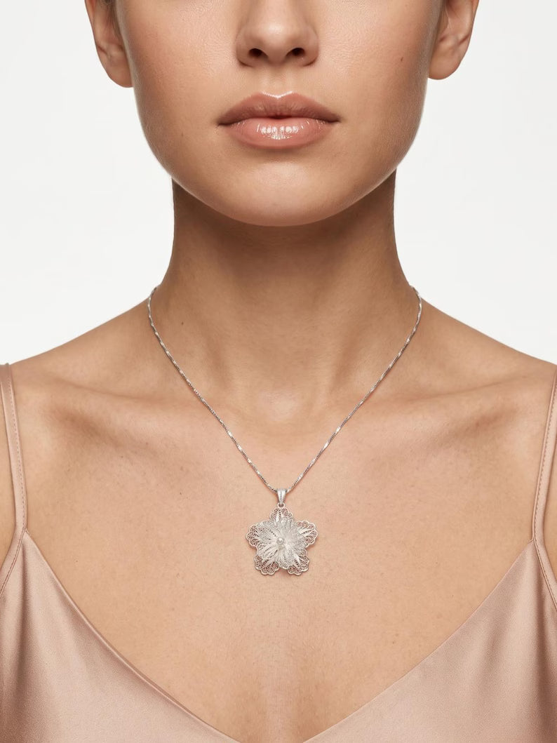 A close-up of a model wearing a delicate silver chain necklace with a textured, flower-like silver pendant against a clean, light gray background.
