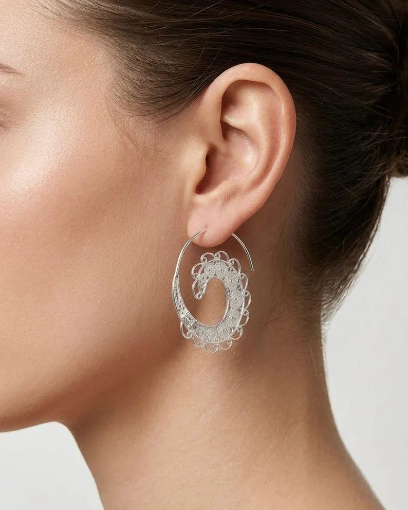 A close-up of a model's ear wearing a large silver hoop earring with an intricate, openwork filigree design.