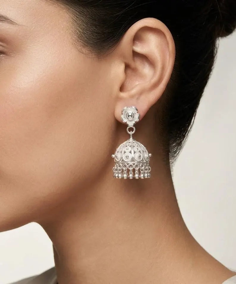 A close-up of a model's ear wearing a silver-toned traditional Indian jhumka earring with intricate filigree detailing and dangling bead chains.
