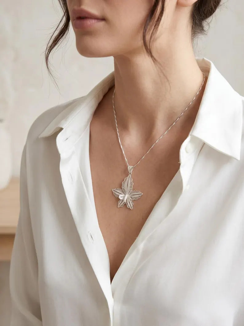 A close-up of a woman wearing a delicate silver necklace with a textured leaf pendant, styled with a white collared blouse.