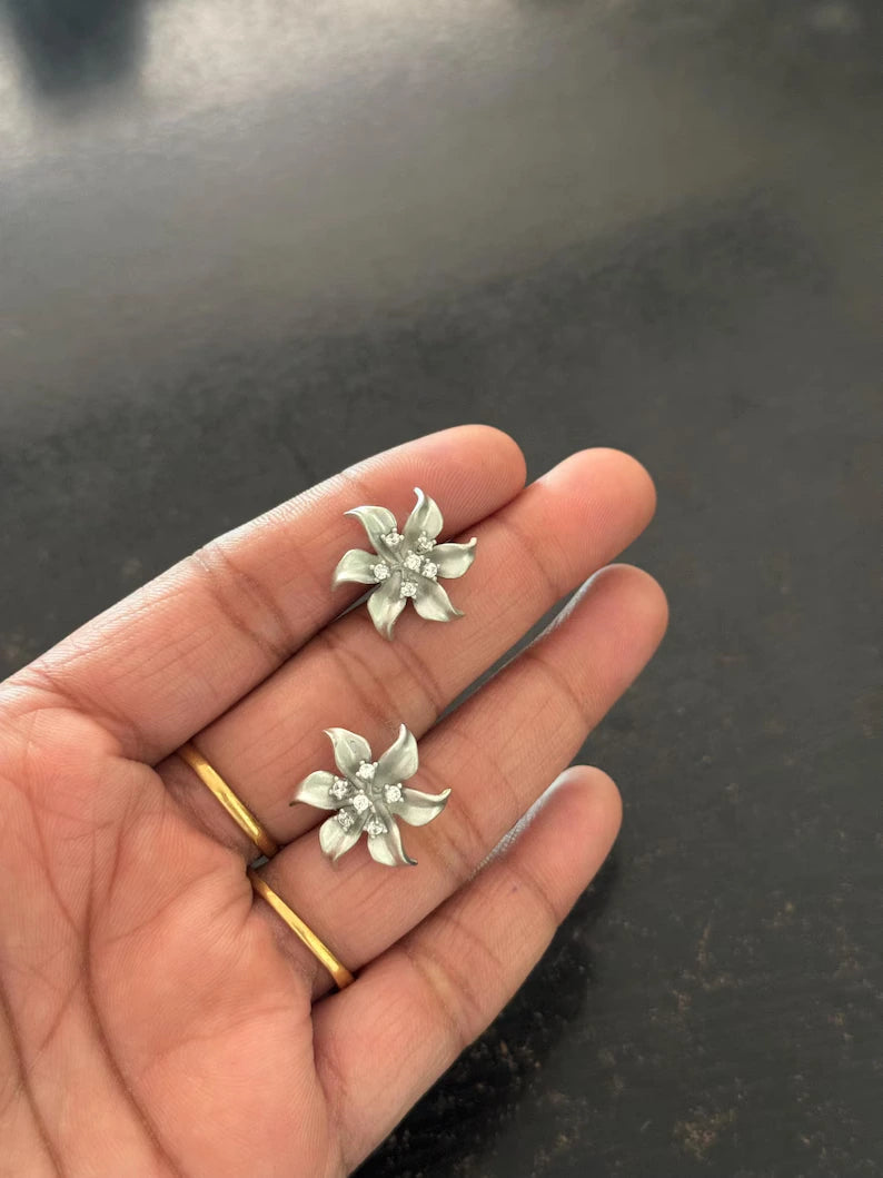 A pair of silver flower-shaped stud earrings held on a person's fingers against a dark gray background.