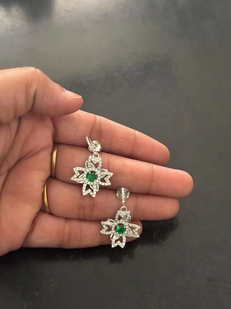 A hand holding a pair of silver-toned dangle earrings with green center stones and clear surrounding stones, shown against a dark gray background.