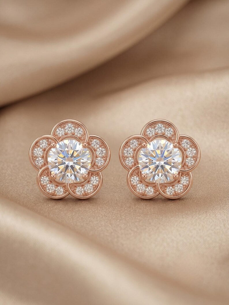 A pair of rose gold flower-shaped stud earrings with clear round-cut center stones and smaller surrounding stones, displayed on a beige satin fabric background.