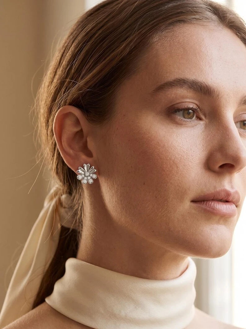 A close-up of a woman wearing a silver flower-shaped stud earring with clear crystals, paired with a cream turtleneck top.